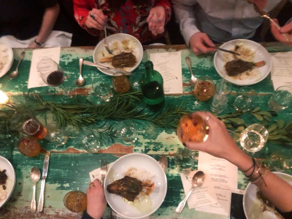 Flatlay photo of rustic green, wooden table with plates of food and diners around it.